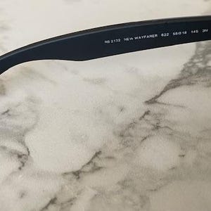 May include: Close-up of a pair of black sunglasses. The arm of the glasses displays the text "RB 2132 NEW WAYFARER 422 55018 145 3N". The lenses are dark green. The glasses are resting on a white and gray marble surface.