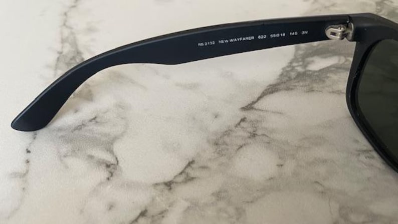May include: Close-up of a pair of black sunglasses. The arm of the glasses displays the text "RB 2132 NEW WAYFARER 422 55018 145 3N". The lenses are dark green. The glasses are resting on a white and gray marble surface.