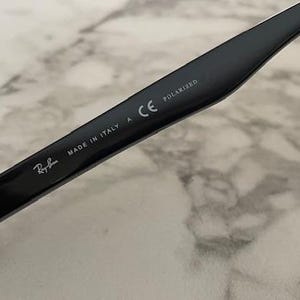 Puede incluir: Primer plano de una patilla de gafas de sol negra con el logotipo de Ray-Ban, texto "MADE IN ITALY" y "POLARIZED". La patilla es negra brillante con bisagras plateadas. Fondo con patr&oacute;n de m&aacute;rmol claro.