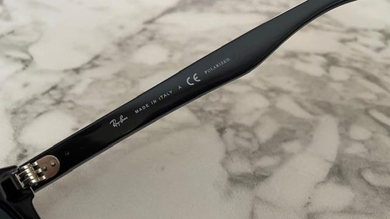Puede incluir: Primer plano de una patilla de gafas de sol negra con el logotipo de Ray-Ban, texto "MADE IN ITALY" y "POLARIZED". La patilla es negra brillante con bisagras plateadas. Fondo con patr&oacute;n de m&aacute;rmol claro.