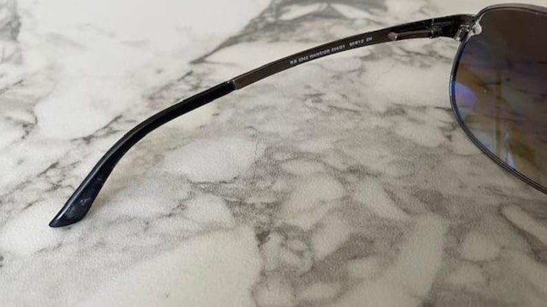 May include: Close-up of sunglasses with a dark frame and gradient lenses. The temple arm is black with text that reads "RB 3045 WARRIOR 204/51 60[]12 2N". The sunglasses are resting on a marble surface.