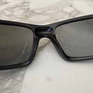 May include: Black sunglasses with rectangular lenses. The sunglasses have a thick black frame and dark lenses. The lenses have scratches. The sunglasses are resting on a white surface.