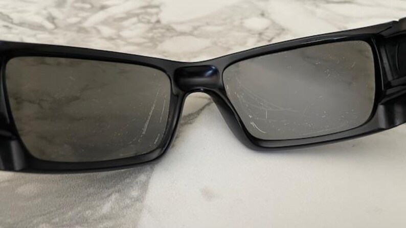May include: Black sunglasses with rectangular lenses. The sunglasses have a thick black frame and dark lenses. The lenses have scratches. The sunglasses are resting on a white surface.