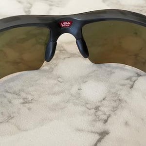 Peut inclure: Lunettes de soleil de sport noires avec des verres teint&eacute;s orange. Les lunettes ont une monture incurv&eacute;e et des plaquettes de nez. Le texte "USA MADE" est visible sur la monture. Ces lunettes sont con&ccedil;ues pour les activit&eacute;s de plein air.