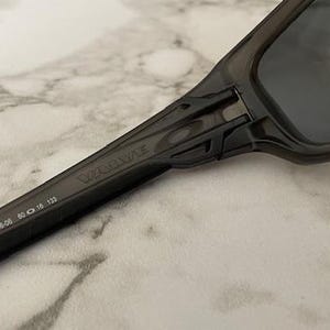 May include: Close-up of dark gray sunglasses with the brand name "Valve" printed on the arm. The arm also displays the numbers "009236-06 60 16 133". The lenses are dark, and the frame is a solid, matte gray.