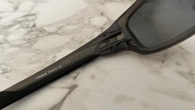 May include: Close-up of dark gray sunglasses with the brand name "Valve" printed on the arm. The arm also displays the numbers "009236-06 60 16 133". The lenses are dark, and the frame is a solid, matte gray.