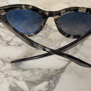 Puede incluir: Gafas de sol con montura de carey y lentes azules. Las patillas de las gafas est&aacute;n cruzadas y las lentes tienen un efecto degradado. Las gafas de sol est&aacute;n sobre un fondo de m&aacute;rmol.