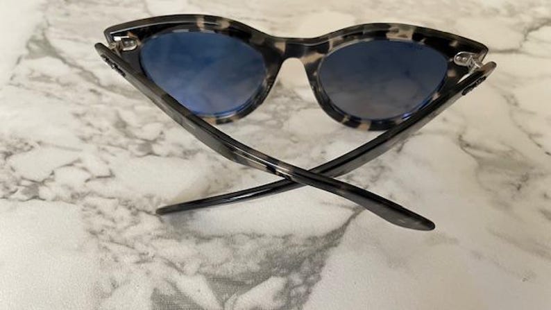 Puede incluir: Gafas de sol con montura de carey y lentes azules. Las patillas de las gafas est&aacute;n cruzadas y las lentes tienen un efecto degradado. Las gafas de sol est&aacute;n sobre un fondo de m&aacute;rmol.