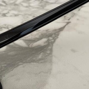 May include: Close-up of black sunglasses with dark lenses. The frame is glossy and rectangular, with a sleek, modern design. The sunglasses are set against a light-colored, marbled surface.