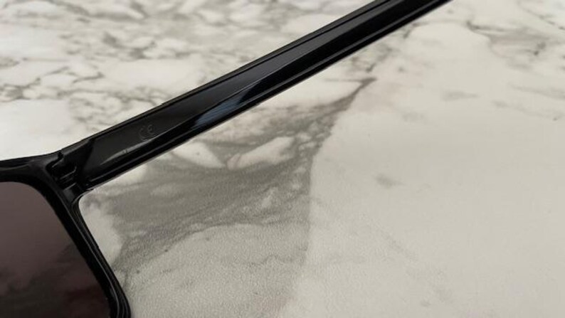 May include: Close-up of black sunglasses with dark lenses. The frame is glossy and rectangular, with a sleek, modern design. The sunglasses are set against a light-colored, marbled surface.