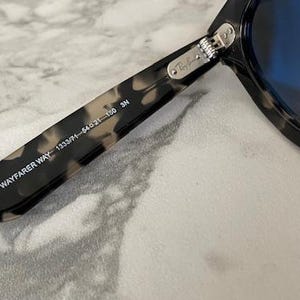 Puede incluir: Primer plano de gafas de sol Wayfarer con montura moteada en negro y marr&oacute;n. Las lentes son de color azul intenso. El texto "RB 2143 WAYFARER WAY 1333/71-54021-150 3N" est&aacute; impreso en la patilla.