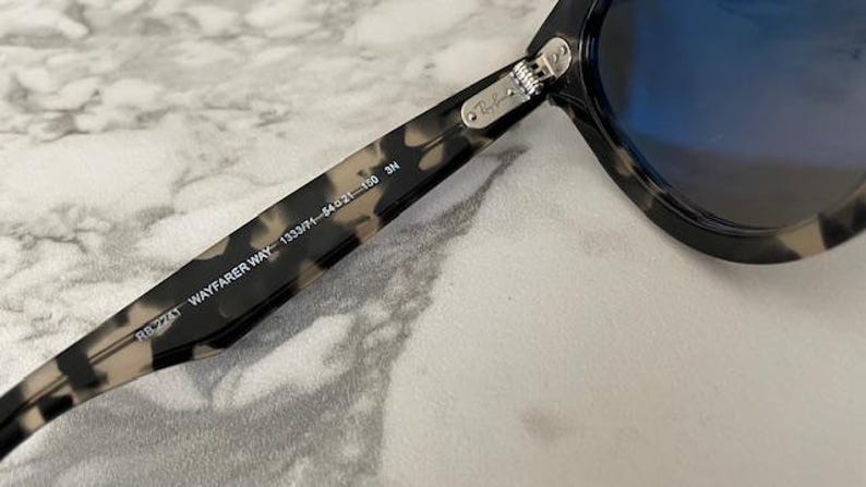 Puede incluir: Primer plano de gafas de sol Wayfarer con montura moteada en negro y marr&oacute;n. Las lentes son de color azul intenso. El texto "RB 2143 WAYFARER WAY 1333/71-54021-150 3N" est&aacute; impreso en la patilla.
