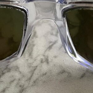 May include: Close-up of clear-framed sunglasses with tinted lenses. The bridge of the glasses is visible, along with the edges of the lenses. The lenses appear to be a dark green color.