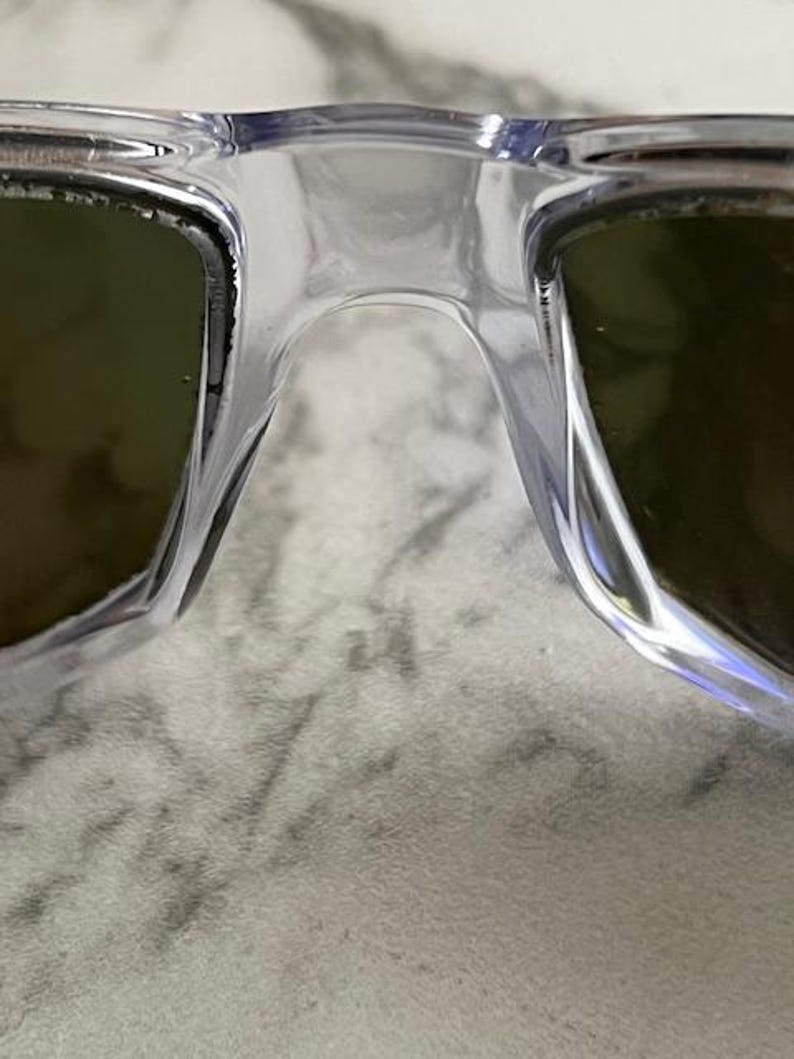 May include: Close-up of clear-framed sunglasses with tinted lenses. The bridge of the glasses is visible, along with the edges of the lenses. The lenses appear to be a dark green color.