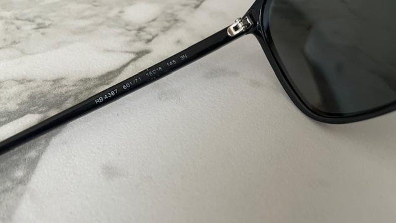 Puede incluir: Gafas de sol negras con dise&ntilde;o de aviador cl&aacute;sico. La montura negra tiene el texto "RB 4387 601/71 56o18 145 3N" impreso en la patilla. Las lentes oscuras contrastan con un fondo claro. Dise&ntilde;o cl&aacute;sico.