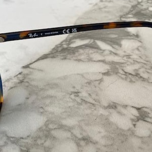May include: Sunglasses with blue lenses and a tortoiseshell pattern on the frames. The arm of the glasses has the brand name and "MADE IN CHINA" printed on it. The glasses are on a marble surface.