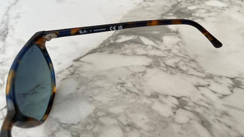 May include: Sunglasses with blue lenses and a tortoiseshell pattern on the frames. The arm of the glasses has the brand name and "MADE IN CHINA" printed on it. The glasses are on a marble surface.