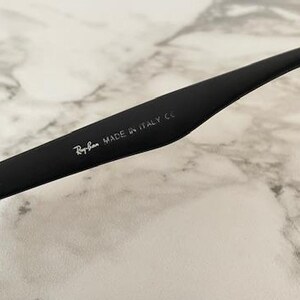 Pu&ograve; includere: Primo piano di un'asta di occhiali da sole nera con il logo Ray-Ban e la scritta "MADE IN ITALY CE" in bianco. L'asta &egrave; curva e ha una finitura opaca. Lo sfondo &egrave; un motivo marmoreo bianco e grigio.