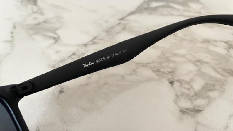 Pu&ograve; includere: Primo piano di un'asta di occhiali da sole nera con il logo Ray-Ban e la scritta "MADE IN ITALY CE" in bianco. L'asta &egrave; curva e ha una finitura opaca. Lo sfondo &egrave; un motivo marmoreo bianco e grigio.