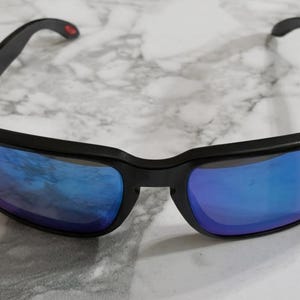 Puede incluir: Gafas de sol negras con lentes azules espejadas. La montura rectangular es negra y las patillas tienen un pequeño detalle rojo cerca de la bisagra.