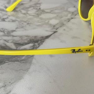 May include: Bright yellow sunglasses with dark lenses. The Ray-Ban logo is visible on the temple. The sunglasses are set against a marble background.