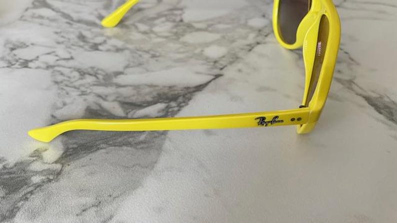 May include: Bright yellow sunglasses with dark lenses. The Ray-Ban logo is visible on the temple. The sunglasses are set against a marble background.