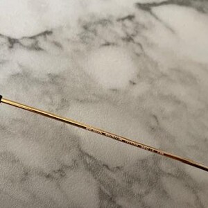 May include: Close-up of a pair of sunglasses. The frame is gold-colored with a black arm. The arm has text printed on it. The background is a light gray and white marble pattern.