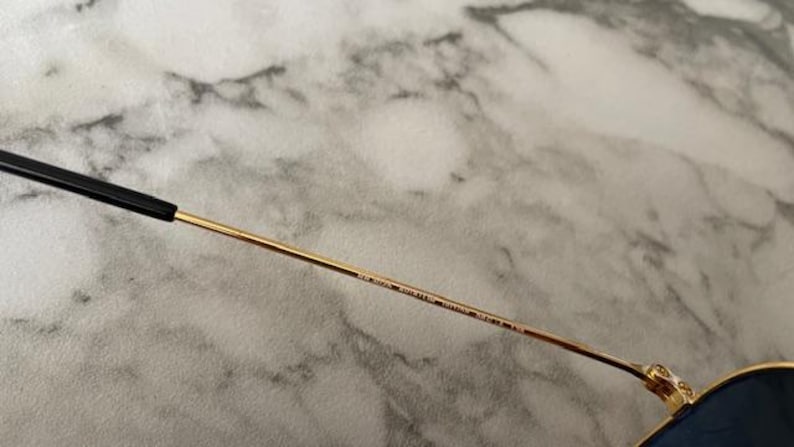 May include: Close-up of a pair of sunglasses. The frame is gold-colored with a black arm. The arm has text printed on it. The background is a light gray and white marble pattern.