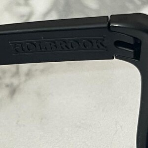 May include: Close-up of a black sunglasses frame with the word "HOLBROOK" embossed on the temple. The sunglasses appear to be made of a matte black material. The image is well-lit, highlighting the details of the frame.