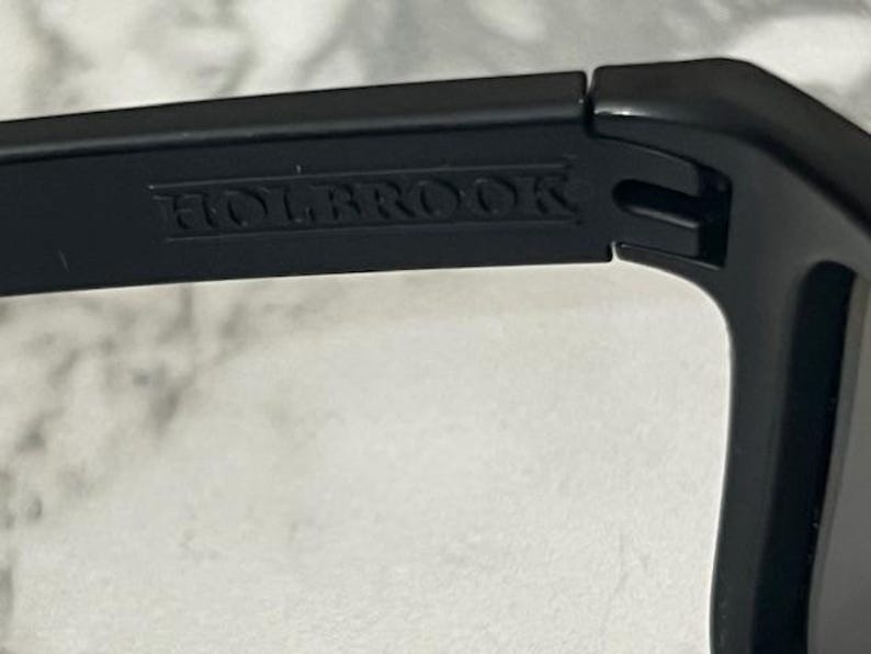 May include: Close-up of a black sunglasses frame with the word "HOLBROOK" embossed on the temple. The sunglasses appear to be made of a matte black material. The image is well-lit, highlighting the details of the frame.