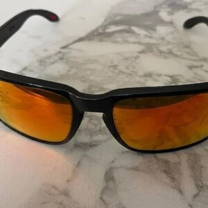 Puede incluir: Gafas de sol negras con lentes de color naranja. La montura rectangular tiene una ligera curva en la parte superior. Pequeños detalles rojos son visibles en la montura. Las lentes reflejan un color naranja brillante.