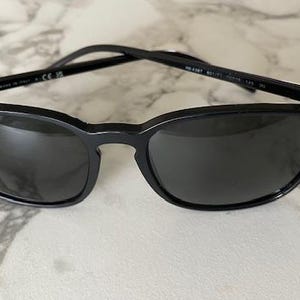 Puede incluir: Gafas de sol negras con lentes oscuras. Las gafas tienen un dise&ntilde;o elegante y moderno con una montura curvada y un peque&ntilde;o logotipo en la lente izquierda. Las patillas son finas y negras. Sobre una superficie de m&aacute;rmol.