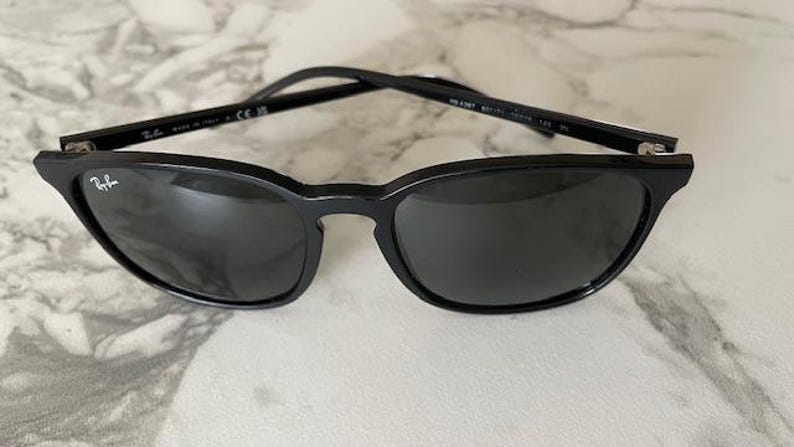 Puede incluir: Gafas de sol negras con lentes oscuras. Las gafas tienen un dise&ntilde;o elegante y moderno con una montura curvada y un peque&ntilde;o logotipo en la lente izquierda. Las patillas son finas y negras. Sobre una superficie de m&aacute;rmol.