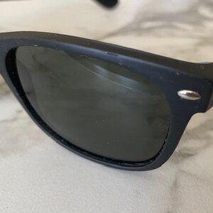 May include: Close-up of black sunglasses with dark lenses. The sunglasses have a classic, rectangular frame with a small silver accent on the temples. The lenses are a dark shade, providing shade from the sun. The sunglasses are a stylish accessory.
