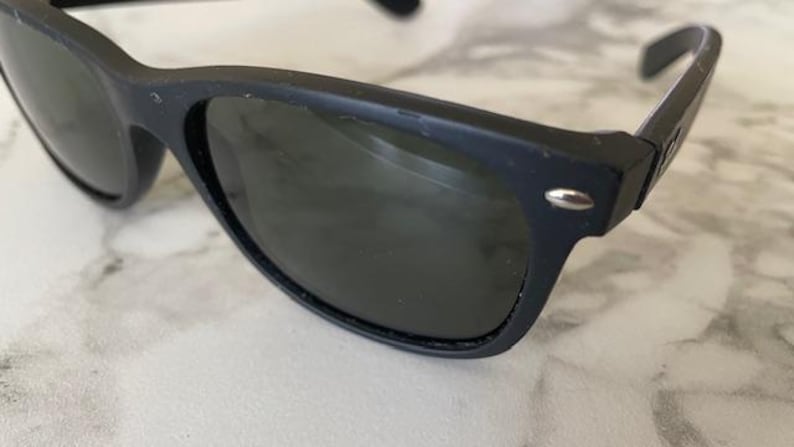 May include: Close-up of black sunglasses with dark lenses. The sunglasses have a classic, rectangular frame with a small silver accent on the temples. The lenses are a dark shade, providing shade from the sun. The sunglasses are a stylish accessory.