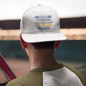 May include: A light gray baseball cap with the text "NURSES CONDEMNING CLOSURE OF USAID KILLING MILLIONS" in blue and yellow. The cap has a flat brim and is worn by a person wearing a green and white shirt.
