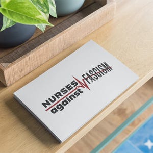 以下が含まれることがあります： 黒と赤で「NURSES against FASCISM」というテキストが書かれた白いカード。単語は赤い心拍ラインで二分されています。カードは明るい木製の表面にあり、鉢植えの植物が入った木製のトレイの近くにあります。