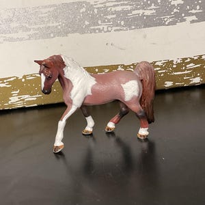 May include: A toy horse figurine in a walking pose. The horse has a white and reddish-brown coat with a white patch on its side. The mane and tail are a darker shade of brown, and the hooves are tan.