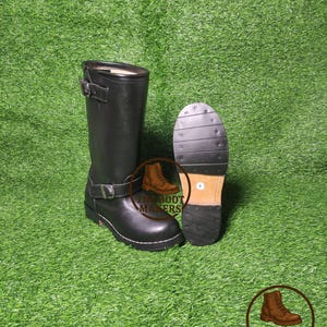 Puede incluir: Botas de moto de cuero negro con hebillas y suela resistente. Una bota está de pie, y la otra se muestra con la suela hacia arriba, mostrando la banda de rodadura. Las botas están sobre un fondo verde.