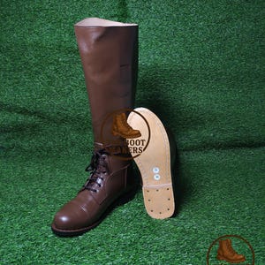 May include: A pair of brown leather combat boots with laces, one upright and the other showing the sole. The boots are on a green surface. The sole is light brown with visible stitching and a size marking. The tall boot is a rich brown color.