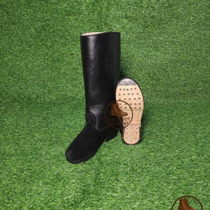 Puede incluir: Bota de montar de cuero negro con empeine de ante y suela en contraste. La bota es alta, llega a la rodilla, y se muestra junto a su suela. La suela es de color marrón claro con un patrón de pequeñas hendiduras circulares. El logotipo de The Boot Makers es visible.