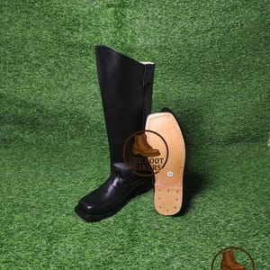 Könnte beinhalten: Schwarze Lederreitstiefel mit quadratischer Spitze und hohem Schaft. Die Sohle eines Stiefels ist sichtbar und zeigt die Nähte und die Konstruktion. Die Stiefel werden auf einer grünen Oberfläche mit dem Logo "THE BOOT MAKERS" präsentiert.