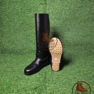 May include: Black leather riding boots with a studded sole, displayed on a green surface. The boots are tall, reaching the mid-calf, and have a classic design. The sole features metal studs for grip. The Boot Makers logo is visible.