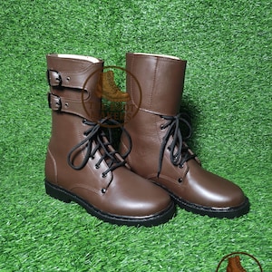 May include: A pair of brown leather boots with black laces and buckles. The boots have a mid-calf height and a sturdy sole. The background is green.