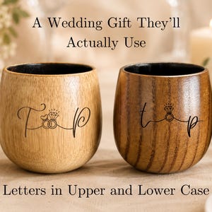 May include: Two wooden cups with black interiors, one light brown and one dark brown. Each cup features a black engraved design of two rings with a diamond and the letters T and P. The text A Wedding Gift They’ll Actually Use and Letters in Upper and Lower Case is visible.