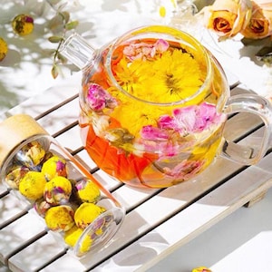 May include: A clear glass teapot and a glass bottle filled with yellow and pink flower buds. The teapot is filled with water and flowers, creating a colorful floral tea. The bottle has a wooden lid.