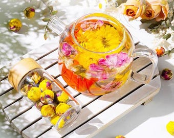 May include: A clear glass teapot and a glass bottle filled with yellow and pink flower buds. The teapot is filled with water and flowers, creating a colorful floral tea. The bottle has a wooden lid.