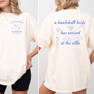 Op de afbeelding: Crèmekleurig T-shirt met blauwe tekst en afbeeldingen. De voorkant leest "Bride Bombshell" met een cocktailillustratie en "ANNA'S BACH Miami Beach, FL." De achterkant zegt "a bombshell bride has arrived at the villa" met schelp-, golf- en vuurtoren-afbeeldingen.