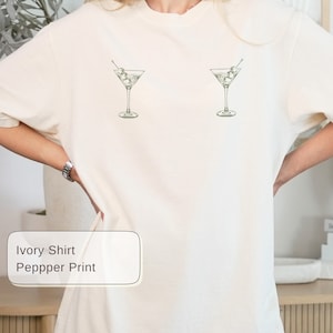 May include: Cream-coloured t-shirt with a minimalist design featuring two martini glasses with olives. The shirt has a relaxed fit and is made of a soft fabric. The text "Ivory Shirt Peppper Print" is in a white rectangle.