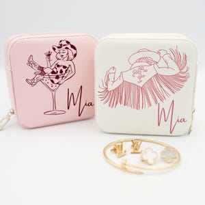 May include: Two square, zipped jewellery cases, one pink and one cream, with illustrations of a cowgirl and the name "Mia". A gold bracelet with charms is in front of the cases. The cowgirl illustrations are in a line drawing style.